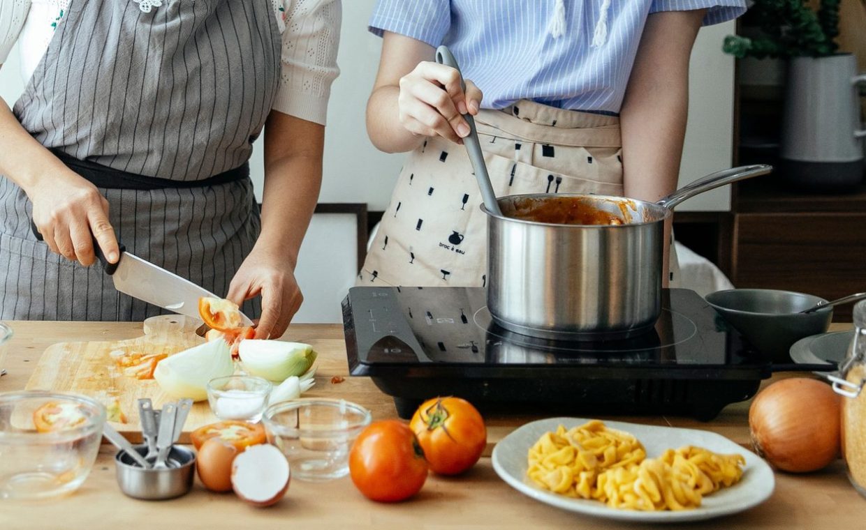 Unrecognizable woman cutting fresh tomato on cutting board while standing at table with ingredients and stove near femal